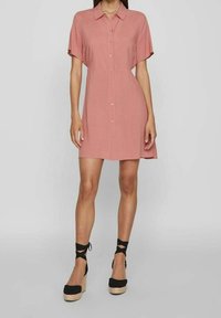 Vestito a maniche corte, con bottoni, in morbido tessuto rosa, caratterizzato da una linea dritta e un retro leggermente più lungo. Abbinato a espadrillas nere con zeppa.