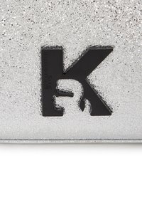 Sidabrinė tekstūruota danga su įtrūkusią paviršiumi, kuriame dominuoja juodas "K" logotipas. Medžiaga atrodo sintetinė su lygiu kraštu.