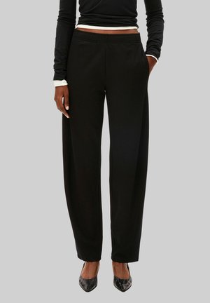 Personne portant un pantalon noir taille haute à jambes larges, un haut noir à manches longues et des chaussures noires à bout pointu, debout devant un fond uni.
