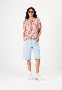 Kortsleeved korallbluse med hvit blomstermønster, parret med lyseblå denim shorts og hvite høyhælte sneakers. Detaljer inkluderer en nøkkelhull-krage.