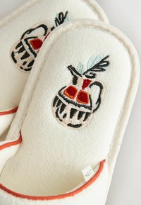 Zapatillas de tela blanca con ribete naranja, cada una con una jarra bordada con patrones decorativos en rojo, negro y azul claro.