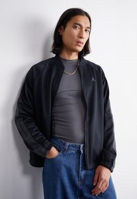 Sort sportsjakke med lynlås, ribbet krave og Jordan-logo. Laget over en tætsiddende grå top og parret med blå jeans.