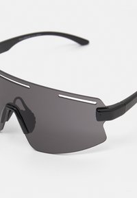Ochelari de soare negri cu lentile mari, curbate și întunecate, având accente subțiri albe în partea de sus și brate negre elegante pentru un design sportiv.