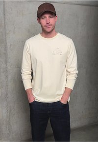 Beige košile s dlouhým rukávem, bílými akcenty a potiskem na levé hrudi, spárovaná s tmavě modrými džíny; jednofázová barva, volný střih.