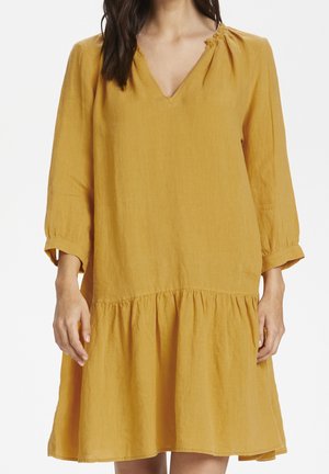 Mujer con vestido amarillo mostaza de manga larga, escote en V y dobladillo fruncido, posando frente a un fondo blanco.