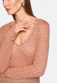 Strickpullover in sanftem Pastellrosa mit strukturiertem Muster und tiefem V-Ausschnitt. Lange Ärmel mit engem Bündchen. Große, runde Ohrringe.