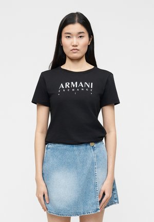 Femeie cu păr lung și închis la culoare, purtând un tricou negru Armani Exchange și o fustă de blugi albastru deschis, stând în picioare într-un decor simplu.