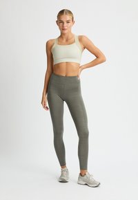 Reggiseno sportivo beige chiaro con spalline larghe, abbinato a leggings verde oliva con un motivo geometrico testurizzato. Scarpe da ginnastica grigie incluse.