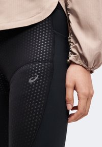 Gros plan d’une personne portant des leggings de sport noirs à pois et une manche de veste beige avec un bouton-pression près du poignet.