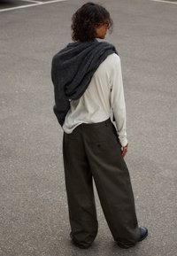 Un pull en laine oversized gris foncé drapé sur une chemise à manches longues claire, associé à un pantalon ample vert olive et des chaussures noires sur l'asphalte.