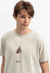 Lichtbeige katoenen t-shirt met een grafische print van een personage in een gestileerde outfit, met korte mouwen en een ronde halslijn.