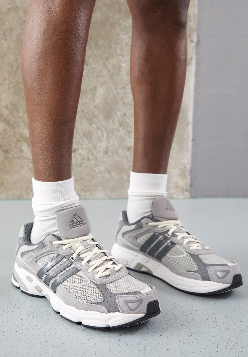 Zapatillas deportivas grises con materiales de malla y ante, que presentan tres rayas gris oscuro, una lengüeta gris acolchada y cordones blancos.