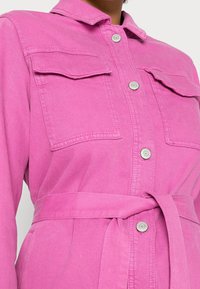Růžová džínová šatová košile s límečkem, dvěma předními kapsami a opaskem v pase. Stříbrné knoflíkové detaily doplňují design.
