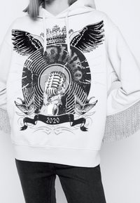 Sudadera blanca con capucha y estampado gráfico que muestra un micrófono, alas y una pancarta con el texto "2020". Flecos decorativos en las mangas.