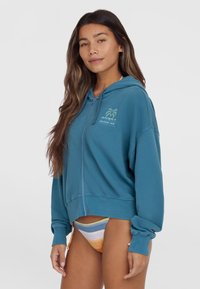 Sweat à capuche zippé teal à coupe décontractée, avec un motif de palmier et des poignets en côtes. Assorti à un bas de bikini rayé orange, bleu et jaune.