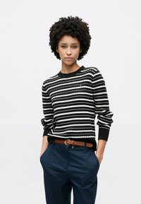 Μπλούζα πλεκτή με ασπρόμαυρες ρίγες και στρογγυλή λαιμόκοψη· συνδυασμένη με μπλε σκούρο παντελόνι και καφέ ζώνη με χρυσή αγκράφα.