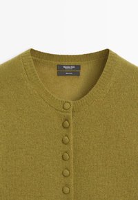 Cardigan in lana verde oliva con scollo rotondo, caratterizzato da sei bottoni abbinati sul davanti e bordi a coste. Etichetta visibile.