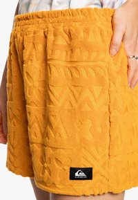 Pomarańczowe szorty o fakturze z wypukłymi kwiatowymi i geometrycznymi wzorami, z elastycznym pasem i małą czarną naszywką z logo.