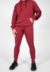 Czerwona bluza z kapturem i czarnymi paskami, w zestawie z dopasowanymi czerwonymi spodniami dresowymi slim-fit. Posiada kieszenie i detale z logo.