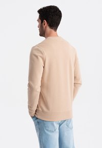 Sudadera beige hecha de tela suave, con cuello redondo, puños y dobladillo acanalados, combinada con jeans de un azul claro. Vista desde la parte posterior.