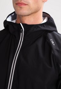 Veste à capuche noire avec une fermeture éclair sur toute la longueur à l'avant, doublure blanche et accents réfléchissants sur les épaules et le logo pour plus de visibilité.