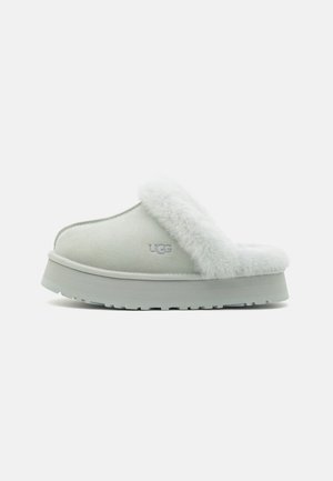 Pantofole grigio chiaro con una morbida fascia in pelliccia, suola piatta e testurizzata, e il logo UGG ricamato in modo discreto sul lato.