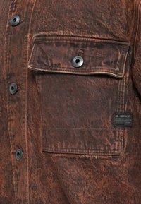 Brown denim shirt with a textured finish, featuring a chest pocket, black snap buttons, and a woven label at the hem.