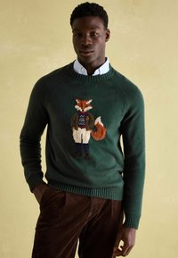 Mann trägt einen grünen Pullover mit gesticktem Fuchs, eine Jacke und einen weißen Hemd, sowie braune Cordhosen, während er vor einem beigen Hintergrund steht.