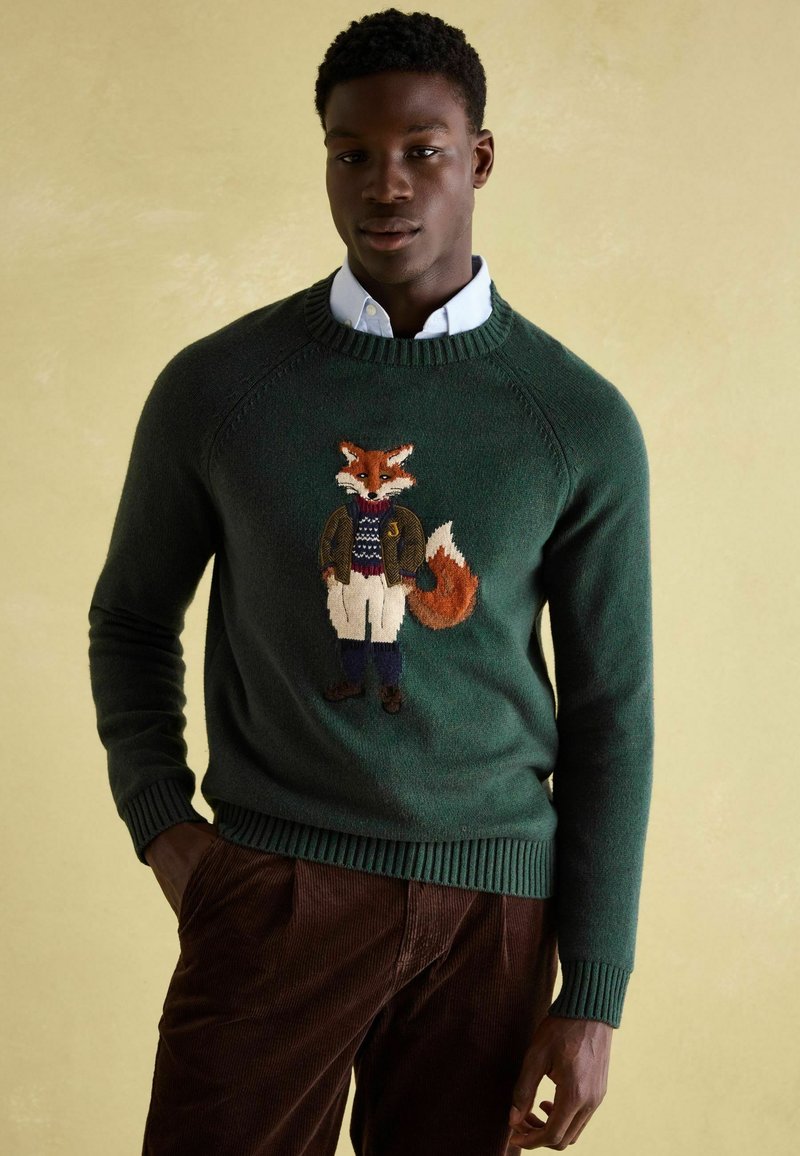 Mann trägt einen grünen Pullover mit gesticktem Fuchs, eine Jacke und einen weißen Hemd, sowie braune Cordhosen, während er vor einem beigen Hintergrund steht.