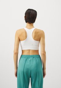 Λευκό αθλητικό στηθόδεσμο με σχέδιο racerback, που διαθέτει εφαρμοστό λάστιχο στη μέση. Συνδυάζεται με φαρδιά, τιρκουάζ αθλητικά παντελόνια.