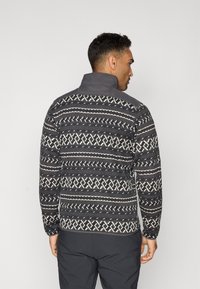 Veste en polaire tricotée de couleur gris foncé avec un motif géométrique crème, un col haut et des manches longues. Présente une texture lisse et des poignets élastiques.