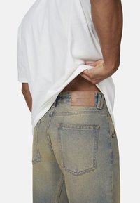 Jean en denim délavé avec une étiquette en cuir sur la ceinture, design à cinq poches et détails de couture visibles.