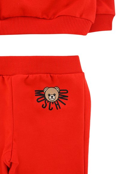 Tuta in cotone rosso con polsini a coste. Presenta una grafica di un orso dei cartoni animati e la scritta "MOSCHINO" stampata in nero sui pantaloni. Texture liscia.