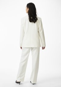 Žena s dlouhými tmavými vlasy, oblečená v krémově bílém obleku s sakem a kalhotami se širokými nohavicemi, stojí zády čelem na bílém pozadí v černých podpatcích.