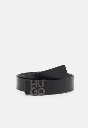 Zwarte leren riem met metalen "HUGO" gesp, opgerold gelegd op een lichtgrijze achtergrond.