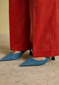 Zilie auduma augstpapēži ar smailiem purngaliem un teksturētu apdari, saskaņoti ar platām sarkana korduroja biksēm. Papēžiem ir neliels melns akcents.