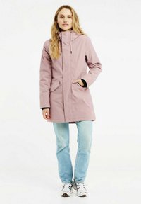 Manteau imperméable rose avec un col haut, fermeture éclair sur le devant et deux poches latérales. Porté sur un haut noir et un jean bleu clair avec des baskets.