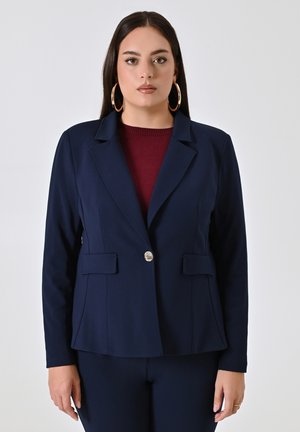Navyblazer aus glattem Stoff, mit einer einzelnen Knopfverschluss, zwei Fronttaschen und einem strukturierten Design. Die Trägerin trägt ein kastanienbraunes Oberteil.