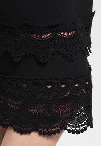 Schwarzer Rock mit aufwendiger Spitze, die gezackte Kanten und florale Muster aufweist, geschichtet mit kontrastierendem strukturiertem Stoff.