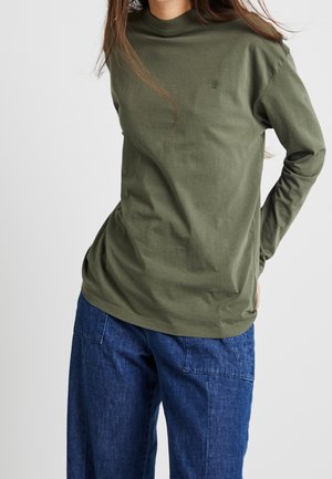 Kobieta ubrana w oliwkowozielony, długi rękaw z golfem oraz luźne niebieskie dżinsy, stojąca z rękami za plecami.