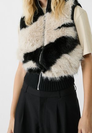 Vrouw draagt een zwart-beige, zachte mouwloze vest met ritssluiting over een crèmekleurige top met korte mouwen, gecombineerd met zwarte broek met hoge taille.