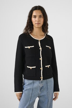 Naine kannab musta tekstuuriga kardigani valge ääre ja kuldsete nööpidega, kombineeritud helesiniste kõrge vöökohaga teksadega, seistes ühevärvilise tausta ees.