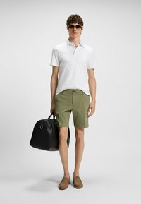 Camisa polo blanca, pantalones cortos olivo, zapatos de lona marrones, bolsa de lona negra, gafas de sol; modelo de pie contra un fondo gris.