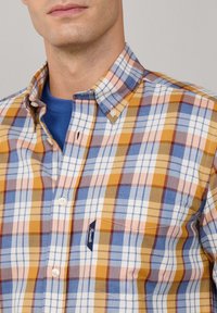 Camisa de cuadros multicolor con tonos azules, amarillos y naranjas. Cuello abotonado y un bolsillo con parche con etiqueta en el pecho. Textura de algodón.