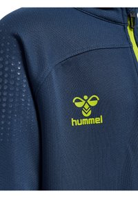Marinblå idrottsjacka med texturerad panel och limegrön dragkedja. Har en liten gul logotyp på bröstområdet. Mjuk tyg med en slät yta.