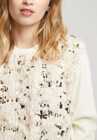 Pull crème avec un devant texturé en tissu moelleux et sequins dorés, contrastant avec un corps en maille lisse.