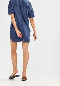 Yksinkertainen denimpuku, jossa on rento malli, lyhyet hihat ja takasauman yksityiskohta. Kangas on tummansininen ja sileä, yhdistettynä mustiin sandaaleihin.