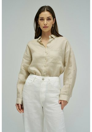 Mujer que lleva una camisa beige abotonada y pantalones blancos de talle alto, posando frente a un fondo liso de color gris claro.