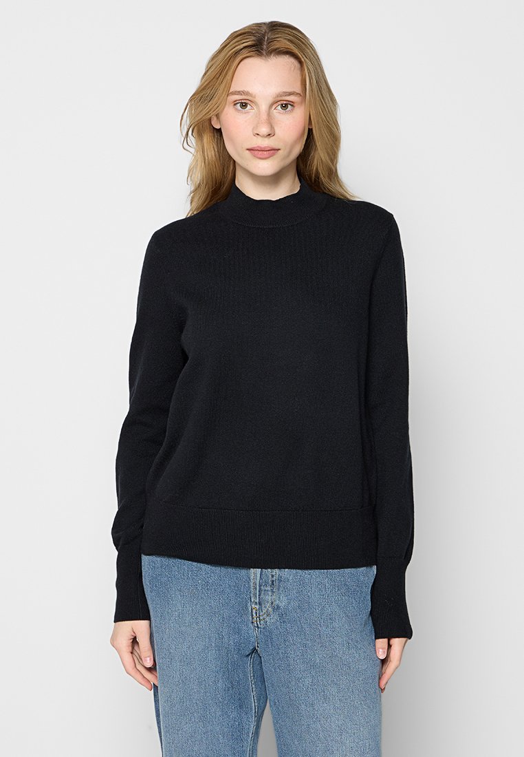 Schwarzer Pullover mit gerippter Textur, hohem Kragen und langen Ärmeln. Hat einen figurbetonten Saum, kombiniert mit hellblauen Jeans.