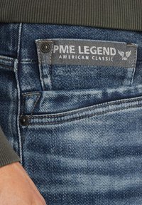 Vrvasti jeans v zbledi modri barvi z teksturirano površino, z sivim oznako iz blaga, ki pravi "PME Legend American Classic."